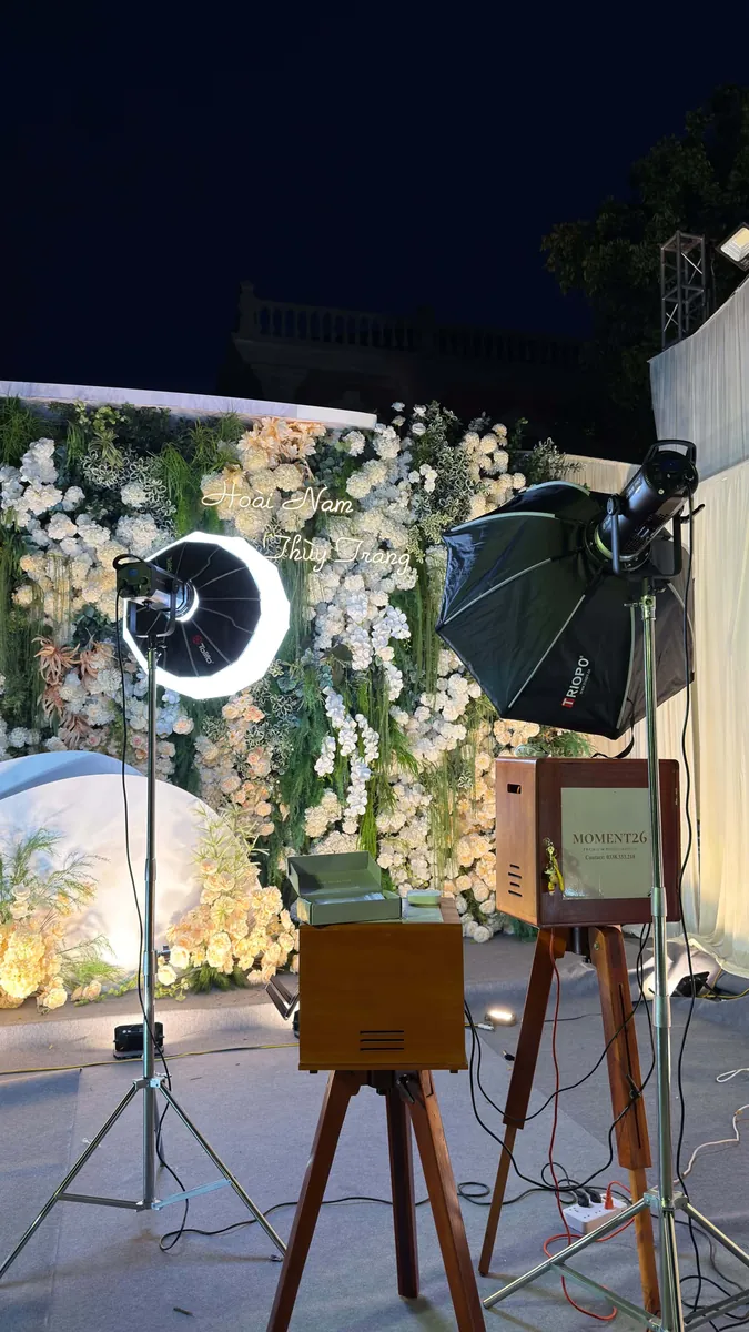 Booth gỗ và đèn studio dựng ngoài trời trước cổng hoa trong tiệc cưới buổi tối